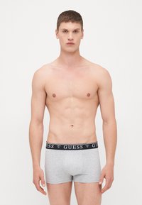 Boxers gris en tissu stretch avec une ceinture élastique noire portant le logo "GUESS". Le tissu semble lisse et extensible. Design à jambe courte.