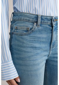 Svetlo modre kavbojske hlače, oblečene z modro-belo ogrnjeno srajco z dolgimi rokavi, prikazani so detajli sprednjega žepa in zanke za pas.