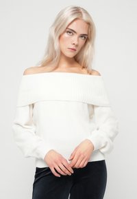 Λευκό πουλόβερ σε στυλ off-the-shoulder με ριγωτή λαιμόκοψη, μαλακή υφή και μακριά, ελαφρώς φουσκωτά μανίκια. Διαθέτει εφαρμοστό ριγωτό στρίφωμα.