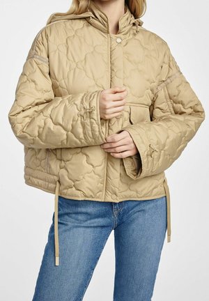 Žena v béžové prošívané bundě s kapucí a modrých džínách, ruce sevřené u přední části bundy u zipu a knoflíků.