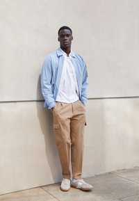 Hellblaues Jackett, weißes Hemd, beigefarbene Cargo-Hose und graue Sneakers mit weißen Akzenten. Glatte Texturen und lässige Designelemente.