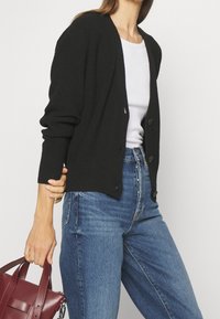 Femme portant un cardigan noir sur un haut blanc et un jean bleu, tenant un petit sac à main rouge foncé dans sa main gauche.