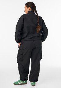 Black oversized jacket with elastic cuffs, paired with black cargo pants featuring side pockets and green sneakers with white stripes.