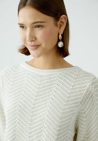 Weißer, gestrickter Pullover mit Zickzackmuster, breitem Ausschnitt und strukturierter Oberfläche. Goldene Tropfenohrringe mit großen Perlen.