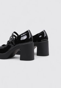 Tacones de charol negro con una plataforma negra gruesa. Presentan una puntera redondeada y una correa decorativa con una hebilla circular.