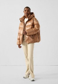 Chaqueta acolchada marrón brillante con capucha y cierre frontal de cremallera. Se lleva con pantalones claros y zapatillas plateadas. Presenta un diseño ajustado y una textura acolchada.