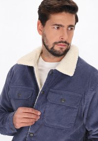 Tengerészkék kordbársony dzseki krémszínű sherpa gallérral, két átfedő zsebbel és sötét gombokkal, texturált anyaggal és laza dizájnnal.