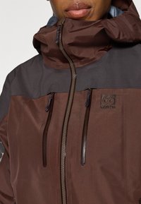 Giacca marrone e grigio scuro con colletto alto, chiusura con zip e due tasche sul petto. Presenta un logo testurizzato sul lato sinistro.