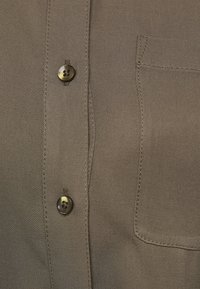 Dettaglio ravvicinato di una camicia in tessuto marrone con due bottoni rotondi scuri e una tasca cucita sul lato destro.
