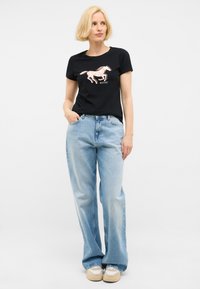 Camiseta negra con un gráfico de un caballo en rosa y texto "MUSTANG". Combinada con unos jeans de campana azules y zapatillas beige con detalles texturizados.