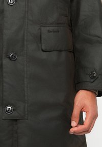 Primeiro plano de uma jaqueta Barbour verde escura com fecho de botões e bolso com aba, mostrando a mão de uma pessoa na manga.