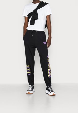 Persoon in zwarte trainingsbroek met paarse en gele tekstdetails, witte t-shirt, zwarte sweatshirt om de schouders gebonden, en zwarte hoge sneakers.