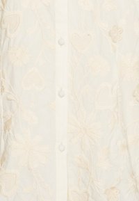 Blusa ricamata color crema con motivi floreali e a cuore. Presenta chiusura con bottoni e un tessuto morbido e textured. Design elegante e dettagliato.