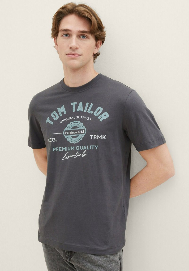 Tricou din bumbac gri cu text imprimat albastru care spune "TOM TAILOR," "CALITATE PREMIUM" și "Esentiale," cu guler rotund și mâneci scurte.