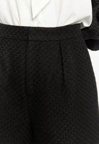 Pantaloni neri texture con un motivo a diamante, caratterizzati da un alto punto vita e pieghe frontali, abbinati a una blusa bianca.