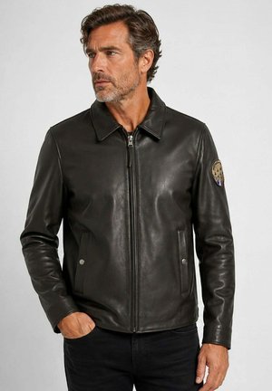 Homme aux cheveux gris courts et à la barbe porte une veste en cuir noire avec une fermeture éclair et un patch sur la manche supérieure, regardant vers la gauche.