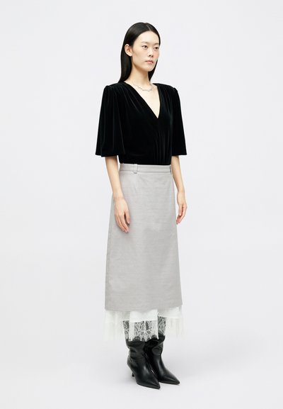 Γυναίκα φορώντας ένα μαύρο βελούδινο τοπ με V-λαιμόκοψη, ανοιχτό γκρι φούστα με λευκό δαντελένιο στρίφωμα, μαύρες μυτερές μπότες και ένα λεπτό κολιέ, στέκεται σε άσπρο φόντο.