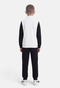 Αγόρι που στέκεται γυρισμένο από την πλάτη, φορώντας άσπρο φανελάκι με μαύρα μακριά μανίκια, μαύρο παντελόνι και άσπρα αθλητικά παπούτσια με μαύρες ρίγες.