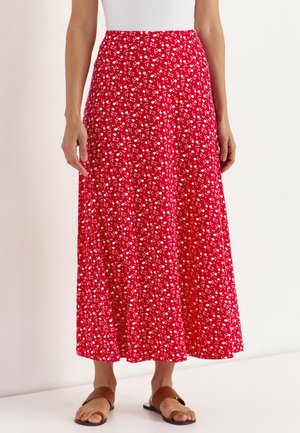 Vrouw draagt een rode midirok met een kleine witte bloemenprint, een wit topje en bruine platte sandalen, staand tegen een eenvoudige lichte achtergrond.