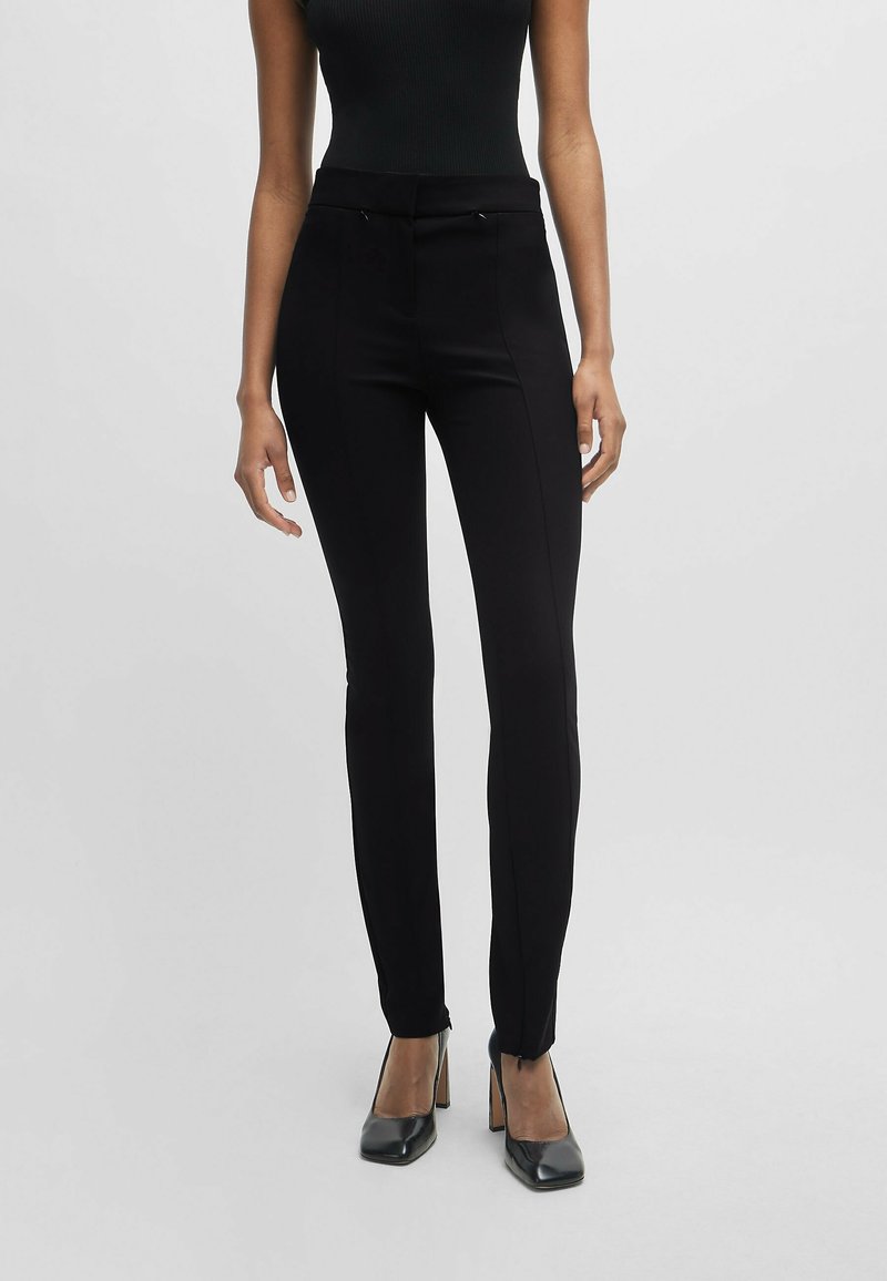 Femme portant un pantalon noir slim et des escarpins noirs, debout devant un fond gris clair.