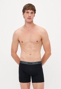 Boxeri scurți negri din bumbac, cu un elastic gri purtând logo-ul "BOSS" în negru. Textură netedă și design ajustat.