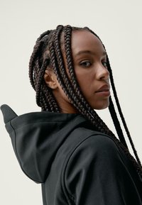 Črna kapucna majica z gladko teksturo, ki ima posebne ramenske šive in pletenice, ki padajo čez eno ramo.