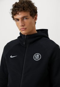 Sort zip-up hættetrøje lavet af en blanding af bomuld; har et logo foran med en løve, en hvid Nike swoosh og teksturerede syninger.
