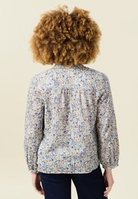 Blusa floreale con maniche lunghe, realizzata in un tessuto leggero con motivi crema, blu e marrone. Dettaglio plissettato sulla schiena.