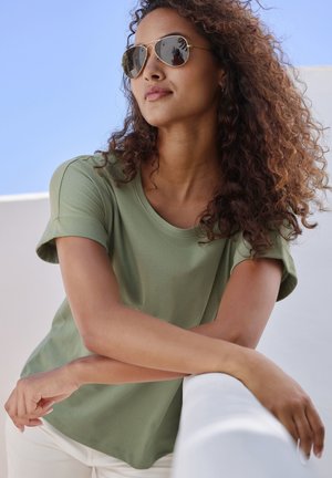 Olivgrünes kurzärmliges T-Shirt mit rundem Ausschnitt, weichem Stoff, kombiniert mit reflektierenden Aviator-Sonnenbrillen vor einem blauen Himmelshintergrund.