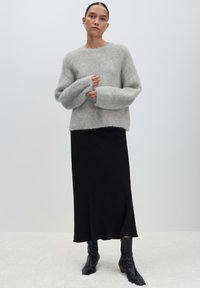 Vrouw in een grijze fuzzy trui, zwarte midi-rok en zwarte leren enkellaarsjes, staande tegen een effen lichte achtergrond.