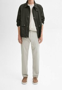 Donkergroene, lichtgewicht jas met drukknoopjes, wit t-shirt, lichtgrijze broek en bruine suède sneakers met witte zolen.