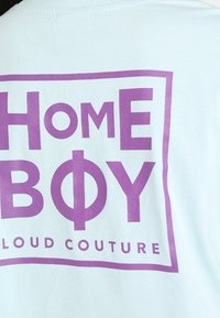 Světle modré bavlněné tričko s velkým fialovým potiskem, na kterém je v tučném písmu napsáno "HOME BOY LOUD COUTURE".