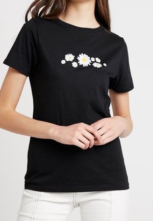 Zwart T-shirt met korte mouwen met witte en gele madeliefjesprint over de borst, gedragen met witte broek.