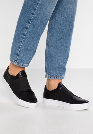 Persoon met een blauwe spijkerbroek en zwarte slip-on sneakers met brede elastische banden en witte zolen, staand op een lichtgekleurde vloer.