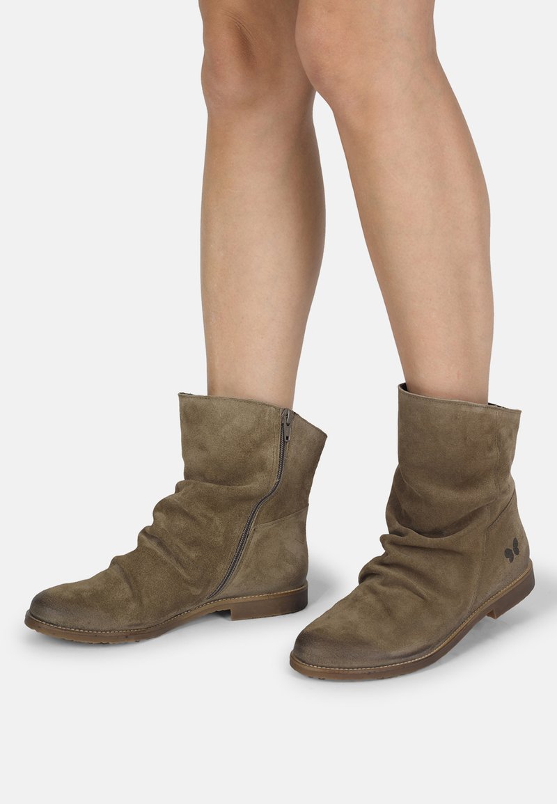 Botas de ante marrón hasta el tobillo con cremalleras laterales, llevadas sobre piernas desnudas contra un fondo blanco.