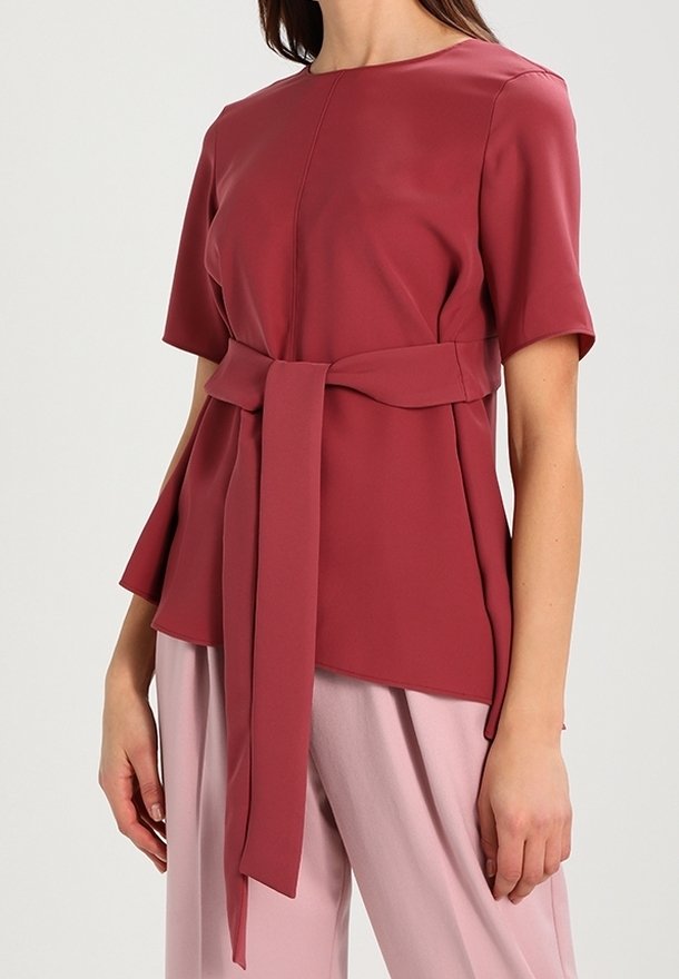 Femme portant un haut rouge foncé à manches courtes avec une large ceinture en tissu nouée à la taille, associé à un pantalon rose clair.