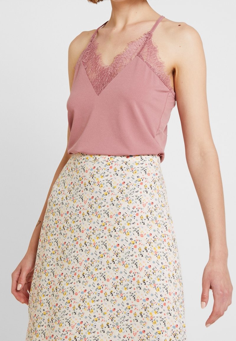Femme portant un haut sans manches rose bordé de dentelle, rentré dans une jupe blanche taille haute avec un petit motif floral multicolore.