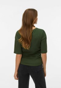 Žalia trumpomis rankovėmis palaidinė, laisvo silueto, su apvalia iškirpte ir švelnia tekstūra, derinama su tamsiomis džinsinėmis kelnėmis. Plaukai krenta žemiau pečių.