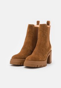 Botas de ante marrón hasta el tobillo con un tacón de plataforma grueso, paneles elásticos a los lados y tiradores para ponérselas fácilmente. Superficie texturizada y suela resistente.