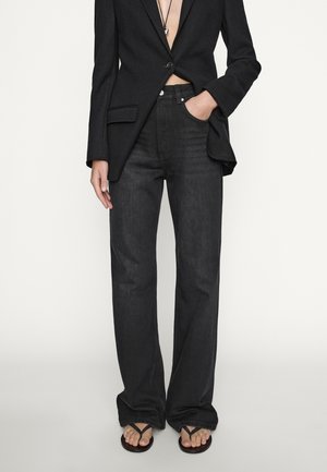 Personne portant un jean noir taille haute à jambes larges, un blazer noir à un bouton, un collier et des tongs noires, debout devant un fond uni.