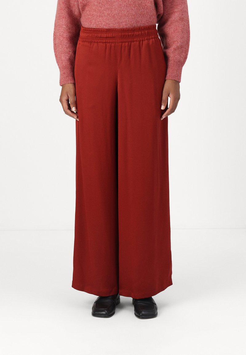 Rode, wijde broek met een elastische tailleband, gemaakt van lichte stof, gecombineerd met zwarte schoenen. Glad oppervlak, minimaal ontwerp.
