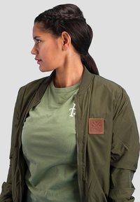 Woman with dark braided hair in a ponytail wearing green jacket with leather patch and matching green t-shirt, looking to her left against gray background.