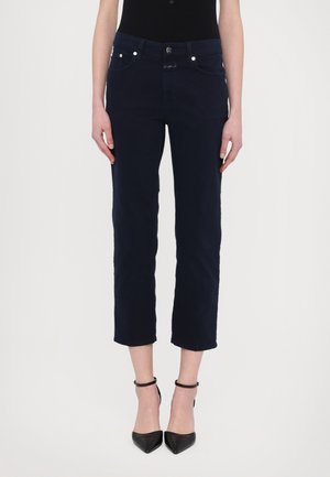 Frau trägt dunkelblaue, verkürzte, gerade geschnittene Hosen im Stil einer Karotte, kombiniert mit schwarzen, spitz zulaufenden Absatzschuhen mit Knöchelriemen, steht vor weißem Hintergrund.