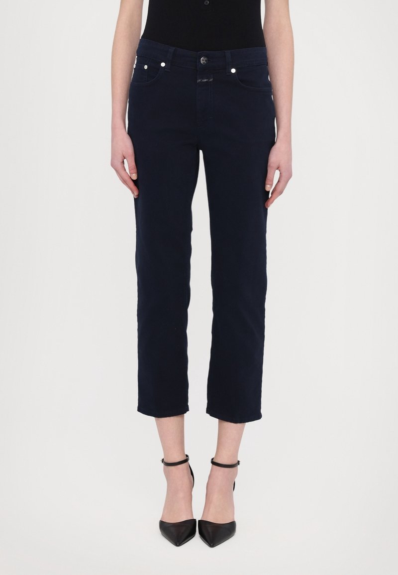 Femme portant un pantalon droit court bleu marine associé à des talons à bride à la cheville noirs à bout pointu, debout devant un fond blanc.