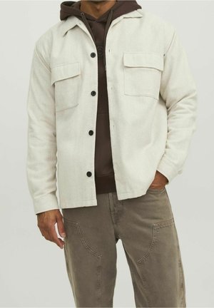 Homme portant une veste beige à boutons par-dessus un sweat à capuche marron foncé et un pantalon taupe avec poches, debout avec une main dans une poche.