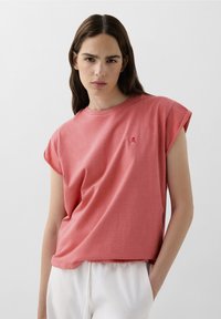 T-shirt corallo a maniche corte con vestibilità morbida, scollatura rotonda e un piccolo logo a forma di teschio nell'angolo in alto a sinistra. Tessuto morbido e strutturato.