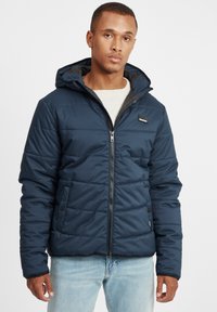 Navy blue padded jacket with a hood, zip closure, and side pockets. Features a smooth texture and a logo patch on the chest.