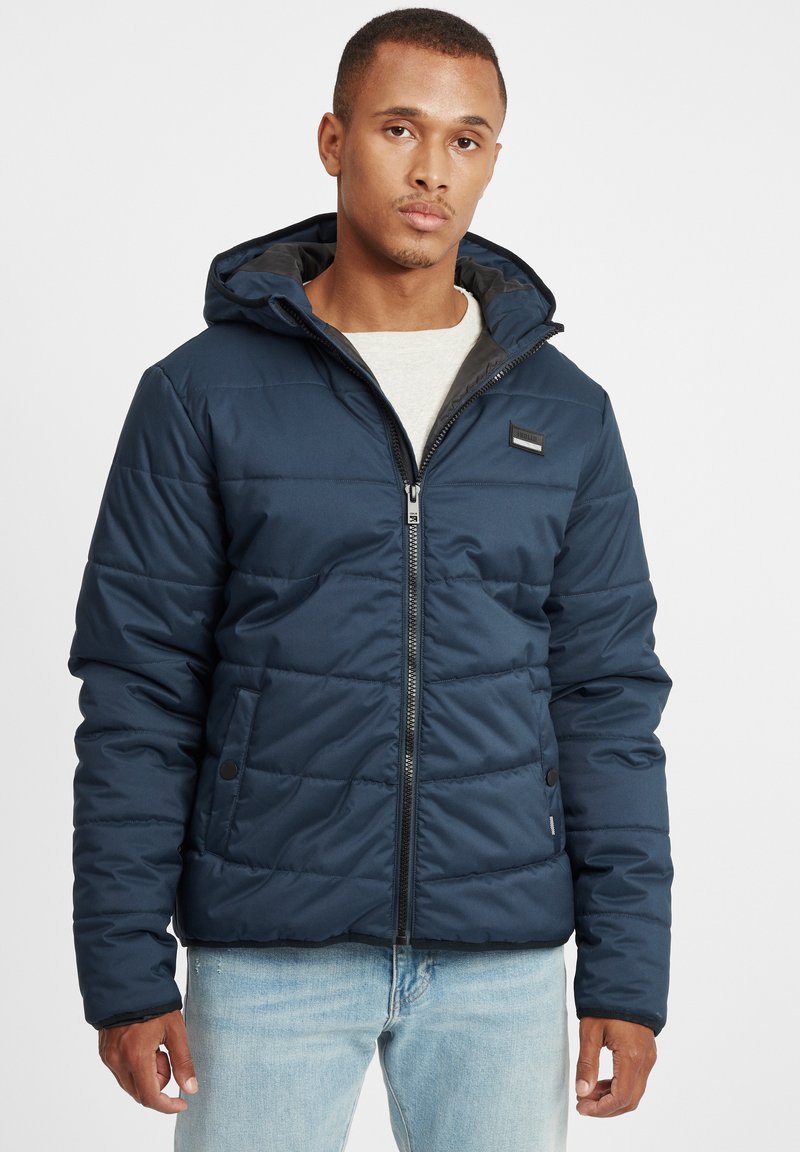 Navy blue padded jacket with a hood, zip closure, and side pockets. Features a smooth texture and a logo patch on the chest.