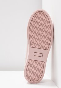 Semelle d'une chaussure rose clair montrant une bande de roulement texturée avec un motif en zigzag et un détail rectangulaire sur une surface blanche et dans un angle de mur.