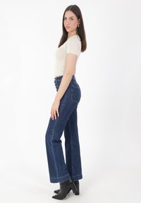 Camiseta ajustada beige combinada con vaqueros acampanados de talle alto en azul oscuro. La modelo lleva botines negros de tacón y está de pie frente a un fondo blanco.